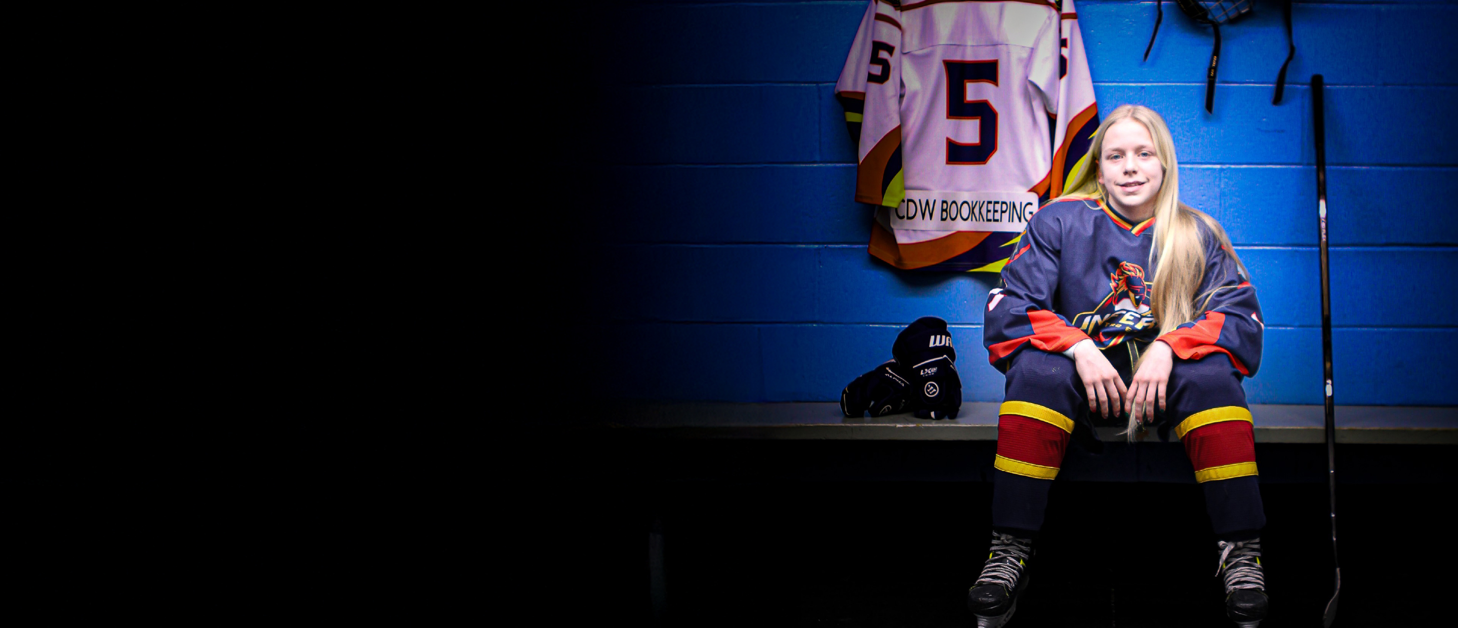 Young hockey player in navy, red, and yellow uniform sitting on a locker-room bench, skates on, with a number 5 jersey labeled “CDW BOOKKEEPING” hanging behind them; hockey gloves and stick nearby against a blue wall.