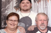 Dakota standing behind his grandparents with his hands on their shoulders, all posing for a family portrait against a rustic wooden backdrop.