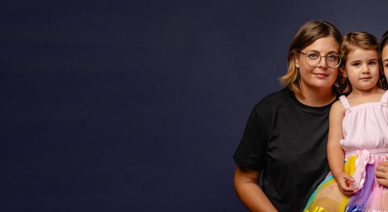 Studio portrait of two women and a toddler smiling together against a dark blue background, modern inclusive family with same-sex parents and young child.