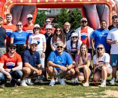 Group of RE/MAX agents posing together outdoors at a community event, wearing branded red, white, and blue shirts in front of colorful inflatable attractions.
